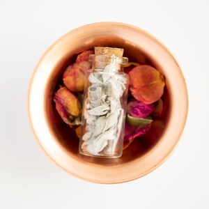 May include: A small glass bottle with a cork stopper filled with dried white sage, sitting in a copper bowl with dried rose petals in shades of pink, red, and orange.