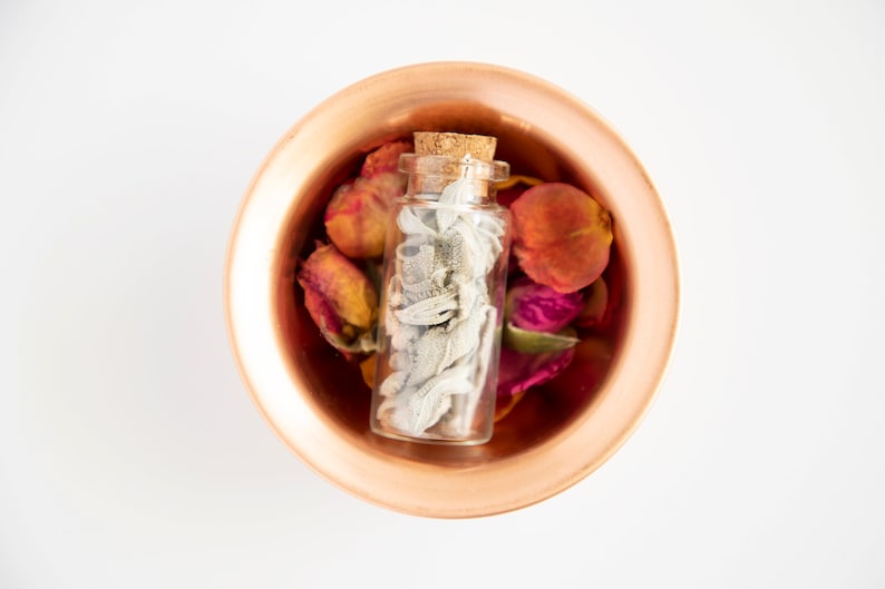 May include: A small glass bottle with a cork stopper filled with dried white sage, sitting in a copper bowl with dried rose petals in shades of pink, red, and orange.