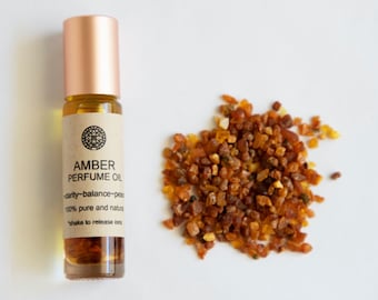 Może przedstawiać: Mała butelka z bursztynowego szkła z korkiem w kolorze różowego złota i brązową etykietą, na której widnieje napis "AMBER PERFUME OIL" z tekstem "clarity-balance-peace" i "100% pure and natural" poniżej. Butelka stoi na białym tle obok kupki bursztynowych kamieni.