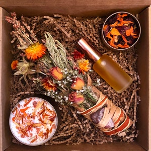 Puede incluir: Una caja de cartón marrón con un ramo de flores secas, un pequeño frasco de flores secas, una botella de vidrio transparente con tapa de cobre y una pequeña vela blanca con flores secas de color naranja y amarillo en su interior.