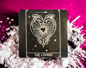 May include: A black tarot card with a white illustration of two figures with wings and a heart in the center. The card reads "THE LOVERS" at the bottom.