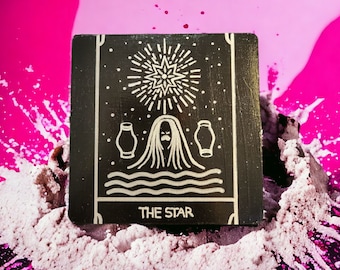 May include: A black tarot card with a white illustration of a star, a figure with flowing hair, and two cups. The card reads "The Star" at the bottom.