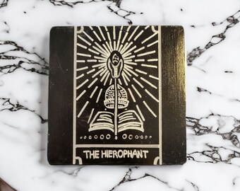 May include: Black wooden coaster with a white design of the Hierophant tarot card. The card features a figure with a staff and a book, surrounded by a radiating sun.