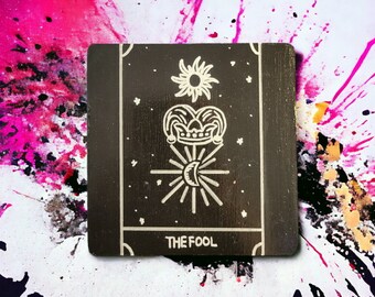 May include: A black tarot card with white illustrations. The card depicts a figure with a crown and a jester's hat, standing on a star with a crescent moon. The text "THE FOOL" is printed at the bottom of the card.