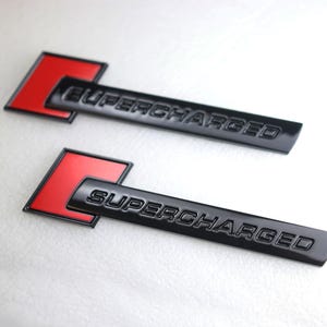 May include: Two black metal car emblems with the word "SUPERCHARGED" embossed in the metal. Each emblem has a red square behind the text.
