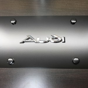 May include: A rectangular black license plate frame featuring the silver metallic Audi logo centered on the front surface. The frame includes four matching black circular screw covers positioned at each corner to secure the plate to a vehicle.