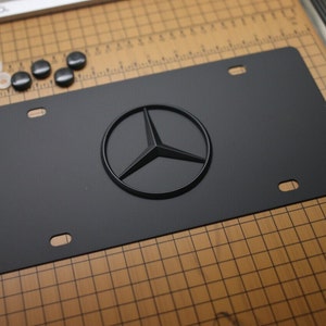 Puede incluir: Placa de matrícula negra mate con el logotipo en relieve de Mercedes-Benz. La placa tiene cuatro orificios de montaje y viene con herrajes negros y blancos. La placa está sobre una superficie cuadriculada.