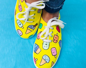May include: A pair of yellow canvas sneakers with white laces, adorned with colorful cartoon ice cream cones and popsicles. The shoes have a white rubber sole and are set against a blue background.