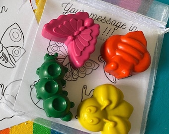 May include: Four shaped wax crayons in pink, orange, green, and yellow inside a sheer white drawstring bag. The bag contains a white card with black line art of insects and the text "Your message in !!!". The crayons are shaped like a butterfly, bee, caterpillar, and flower.