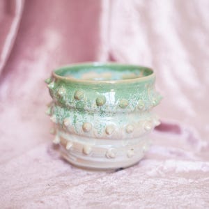 May include: A handcrafted ceramic pot with a textured surface and a unique design. The pot features a light green top that fades into a creamy white base. Small, rounded protrusions encircle the pot, adding a tactile element. The pot is displayed against a soft pink background.