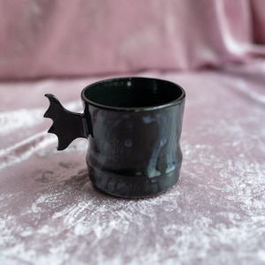 Puede incluir: Una taza de cerámica negra con un asa en forma de ala de murciélago. La taza tiene un acabado brillante y un interior oscuro. El asa es un ala de murciélago estilizada, que añade un toque único y caprichoso. La taza es probablemente hecha a mano.