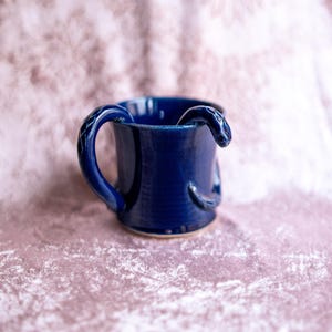 May include: A cobalt blue ceramic mug with a snake design. The handle and the rim of the mug are shaped like a snake. The mug is on a pink surface.