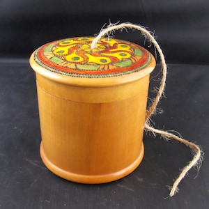 Puede incluir: Una caja redonda de madera clara con tapa decorada con un diseño colorido en amarillo, verde y rojo. La caja es de color marrón claro. Una cuerda de fibra natural está unida a la tapa. Aproximadamente 10 cm de diámetro.