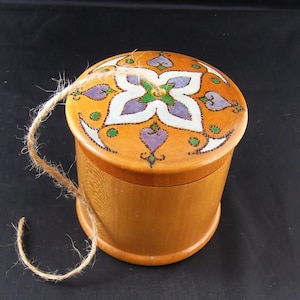 May include: A round wooden box with a lid featuring a floral design in purple, white, and green on an orange background. The box is light brown with a natural wood grain. A length of twine is attached to the lid.