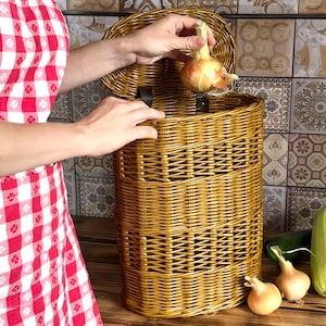 May include: A woven wicker basket with a lid, ideal for storing vegetables. The basket is a warm golden brown color. An onion is being placed inside the basket. Other vegetables are on a wooden surface.
