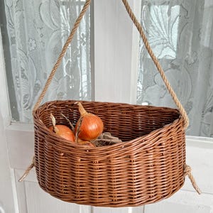 May include: A brown wicker basket with a rope handle, hanging from a hook on a white door. The basket contains two onions. The basket is oval-shaped and has a woven design.