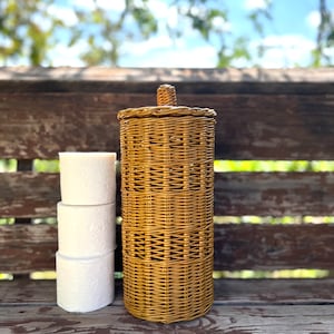 May include: A woven wicker toilet paper holder with a lid. The holder is brown and has a natural finish. Three rolls of toilet paper are stacked next to the holder.