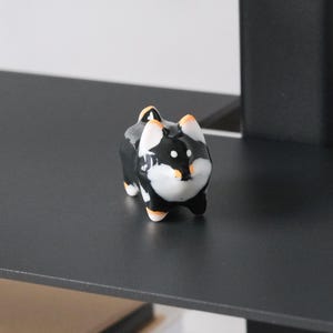 Może przedstawiać: Mała ceramiczna figurka Shiba Inu. Pies jest czarno-biały z pomarańczowymi akcentami na uszach i łapach. Znajduje się na czarnej półce, z książkami poniżej. Figurka to przedmiot dekoracyjny.