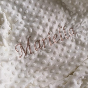 Könnte beinhalten: Weiße Plüsch-Babydecke mit einem erhabenen Punktmuster und dem Namen "Mariella", der in hellbrauner Farbe gestickt ist.