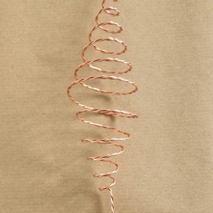 May include: A copper wire sculpture in a spiral form, resembling a Christmas tree or decorative element. The wire is twisted and coiled, creating a three-dimensional design. The background is a solid tan colour.