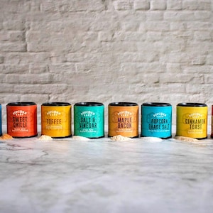 Puede incluir: Ocho botes metálicos de colores con tapas negras y etiquetas blancas. Las etiquetas tienen el texto "TOPCORN" y diferentes nombres de sabores, incluyendo "Chocolate", "Chilli Dulce", "Toffee", "Sal y Vinagre", "Tocino de Arce", "Sal de Palomitas", "Tostada de Canela" y "Dulce y Salado".