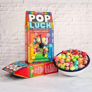 May include: A vibrant box of "Pop Luck" gourmet popcorn dare game and a bowl of assorted popcorn. The box displays the game's title, flavour options, and a clock-like spinner. The popcorn is in various colours, including red, green, and blue.