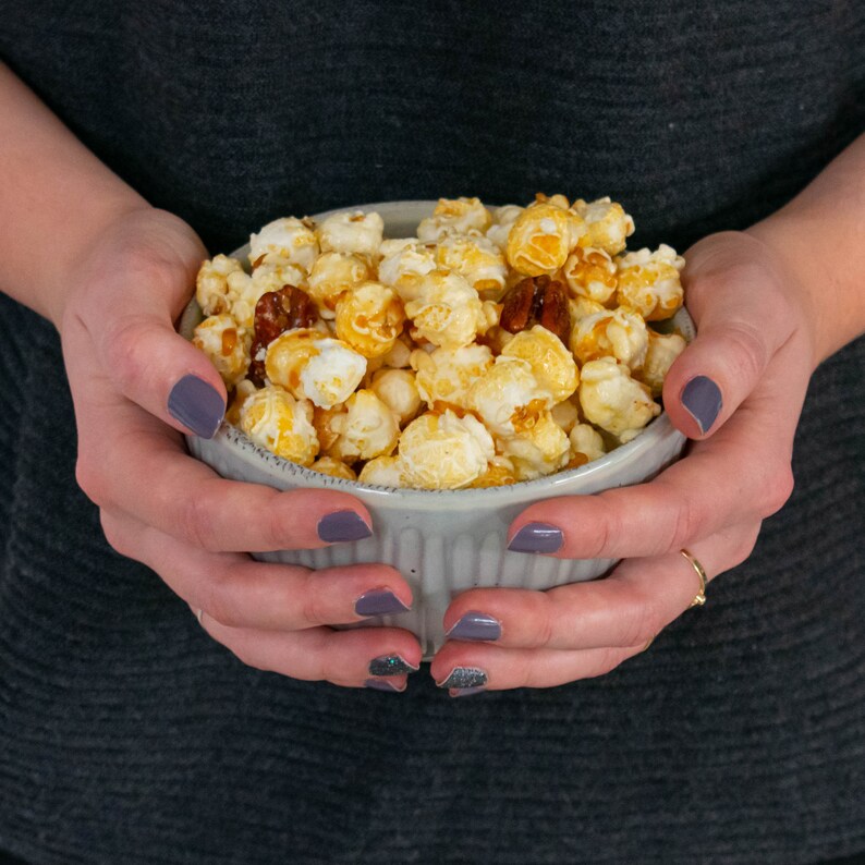 May include: A gray bowl filled with caramel popcorn and pecans. The bowl is being held by two hands.