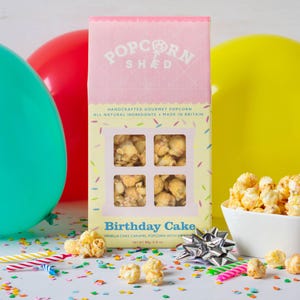 May include: A rectangular box of Popcorn Shed Birthday Cake vanilla cake caramel popcorn with sprinkles. The packaging is pink and yellow with a window showing the popcorn, labeled 80g/2.8 oz, surrounded by colorful balloons and confetti.