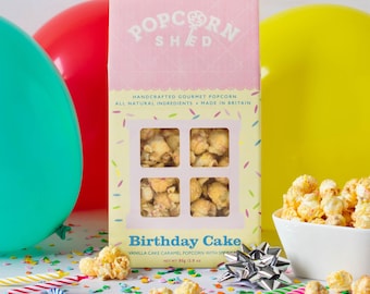 May include: A rectangular box of Popcorn Shed Birthday Cake vanilla cake caramel popcorn with sprinkles. The packaging is pink and yellow with a window showing the popcorn, labeled 80g/2.8 oz, surrounded by colorful balloons and confetti.