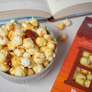 May include: A white bowl filled with caramel popcorn and pecans. The popcorn is drizzled with caramel and has a light, airy texture. The pecans are whole and add a nutty flavor to the popcorn. The bowl is sitting on a white surface.