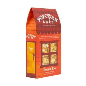 May include: A red and yellow box shaped like a house with a window showing caramel popcorn with pecan pieces. The box says "Popcorn Shed" and "Pecan Pie" on the front. The text also says "Handcrafted Gourmet Popcorn, Gluten Free - All Natural Ingredients".
