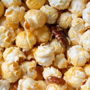 May include: A close-up of a bowl of popcorn with caramel and pecans. The popcorn is a light golden color and the caramel is a rich brown color. The pecans are scattered throughout the popcorn.