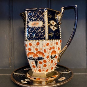May include: A vintage ceramic pitcher with a blue and gold geometric pattern and a white and orange floral pattern. The pitcher has a handle and sits on a matching saucer.
