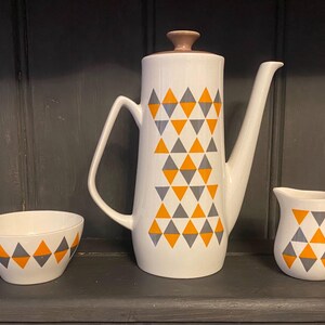 Könnte beinhalten: Eine weiße Teekanne, eine Milchkännchen und eine Zuckerdose aus Keramik mit einem geometrischen Muster aus orangefarbenen und grauen Dreiecken.