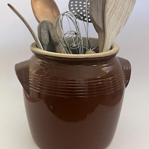 Puede incluir: Un recipiente de cerámica marrón con utensilios de cocina de madera, como cucharas, espátulas y un batidor. El recipiente tiene un diseño acanalado y dos asas laterales. Los utensilios son de diferentes formas y tamaños, creando una estética rústica.
