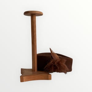 Può includere: Un cappello marrone con una piuma e un porta cappello in legno. Il cappello è di colore marrone scuro e ha una piuma e un'applicazione soffice. Il porta cappello è in legno e ha una parte superiore rotonda.