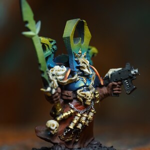 Op de afbeelding: Miniatuurfiguur van een krijger in blauwe, bruine en gouden bepantsering, met een groen zwaard en een zwart pistool. De figuur heeft een schedelachtig helm en is gemonteerd op een zwarte basis. De figuur is ongeveer 5 cm hoog.