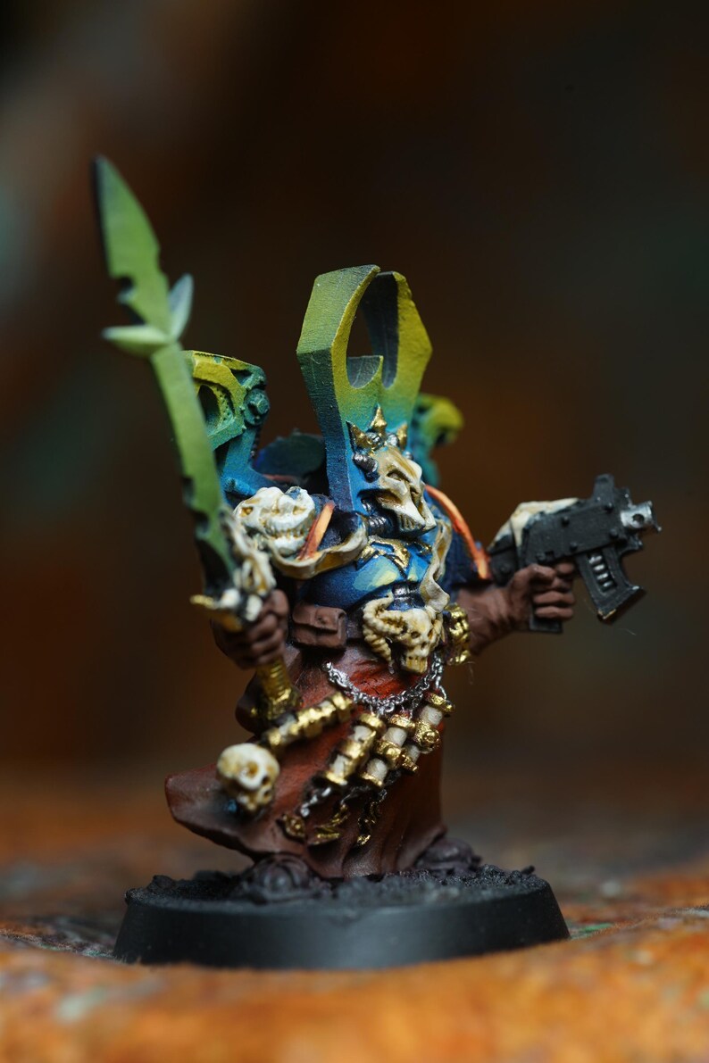 Op de afbeelding: Miniatuurfiguur van een krijger in blauwe, bruine en gouden bepantsering, met een groen zwaard en een zwart pistool. De figuur heeft een schedelachtig helm en is gemonteerd op een zwarte basis. De figuur is ongeveer 5 cm hoog.