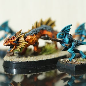 Puede incluir: Figuras de fantasía en miniatura de criaturas reptilianas en tonos de azul, naranja y marrón. Una figura es una bestia grande de cuatro patas con la espalda puntiaguda. Otras figuras son humanoides con lanzas, como en batalla.