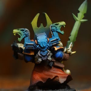 Op de afbeelding: Miniatuurfiguur van een krijger in blauw pantser met groene accenten, met een groen zwaard. De figuur heeft een rode mantel en een geel en groene hoofdtooi. De basis is zwart.
