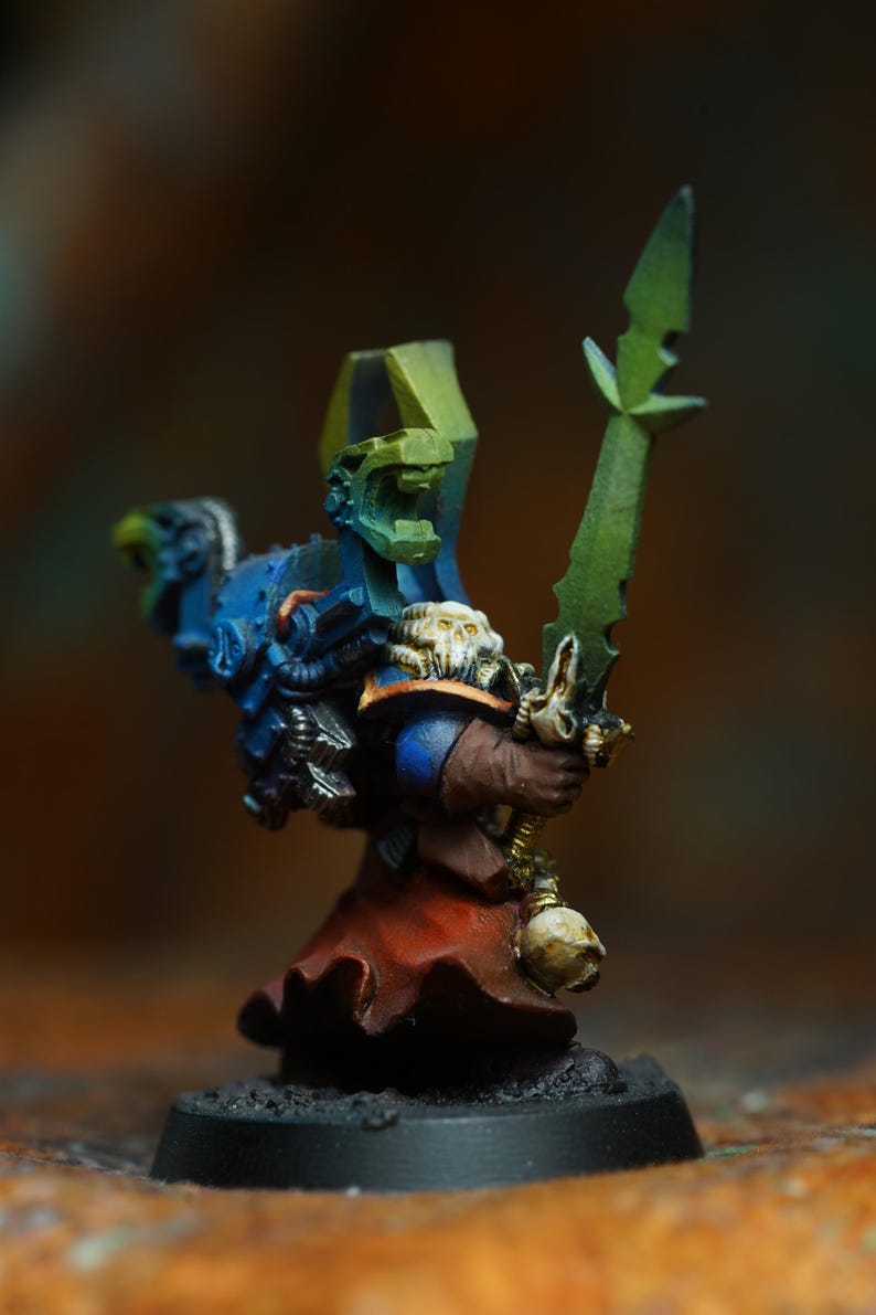 Op de afbeelding: Miniatuurfiguur van een krijger in blauwe, groene en bruine bepantsering, met een groen zwaard. De figuur heeft een met schedels versierde schouderbeschermer en een rode jurk. De basis is zwart.