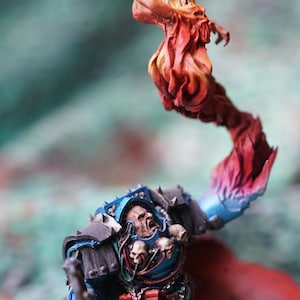 May include: Miniature figurine of a warrior in blue and gray armor, with a red cape and a flaming effect. The figure has a skull-adorned chest and is holding a weapon. The miniature is painted with detailed shading.