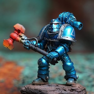 May include: A miniature figurine of a blue armored warrior with a red and orange hammer. The figure is standing on a small, textured rock base. The armor has silver accents and a backpack with a cannon.