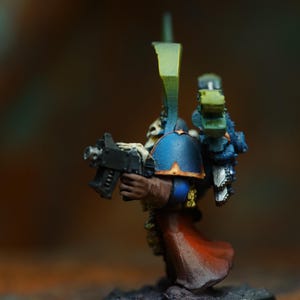 Op de afbeelding: Miniatuurfiguur van een space marine, beschilderd met blauwe, gouden en rode accenten. De figuur houdt een zwart wapen vast en staat op een zwarte basis. De miniatuur is ongeveer 5 cm hoog.