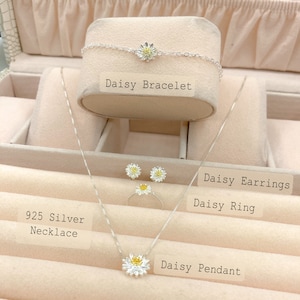 Puede incluir: Un conjunto de joyas de margarita de plata 925, que incluye un collar, una pulsera, pendientes y un anillo. Las joyas se muestran en un joyero con los nombres de cada pieza escritos debajo.