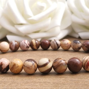May include: A bracelet made of round, natural stone beads in shades of brown, tan, and cream. The beads have unique patterns and colour variations. The bracelet is displayed against a soft background.
