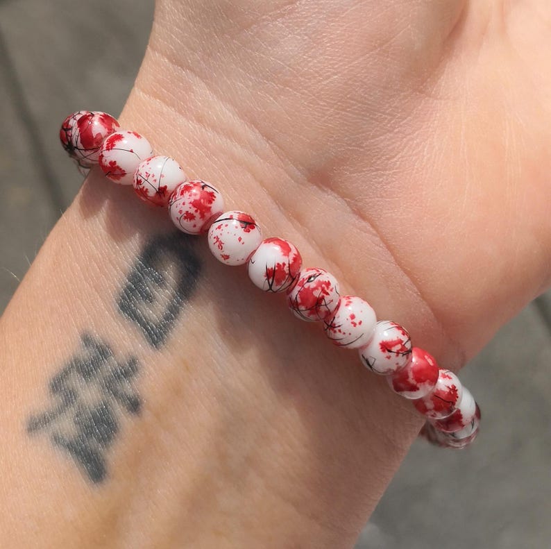 May include: A beaded bracelet featuring round white beads with a red splatter pattern and thin black line accents. The beads are strung together in a single row, creating a circular shape that fits around the wrist. The design is consistent across each individual bead, showcasing a high-contrast color scheme of white, red, and black throughout the entire piece of jewelry.