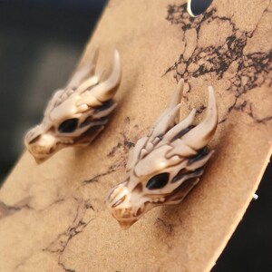 May include: A pair of dragon head earrings made of white resin with black eyes. The earrings are on a brown background.
