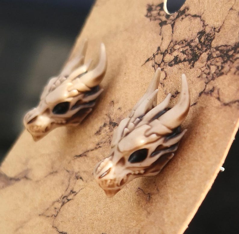 May include: A pair of dragon head earrings made of white resin with black eyes. The earrings are on a brown background.