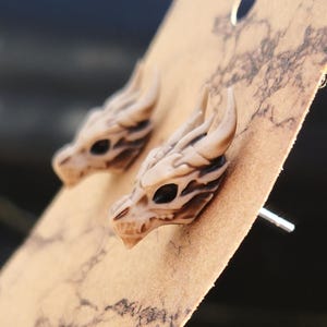 May include: A pair of beige dragon skull stud earrings. The earrings are made of a smooth, textured material and have a detailed design.